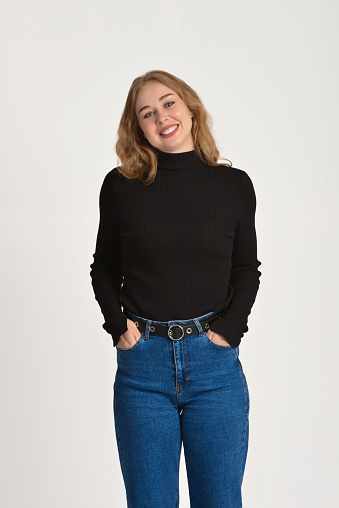 A beautiful smiling young blonde woman stands with her hands in her jeans pockets. Front view. Studio shot.