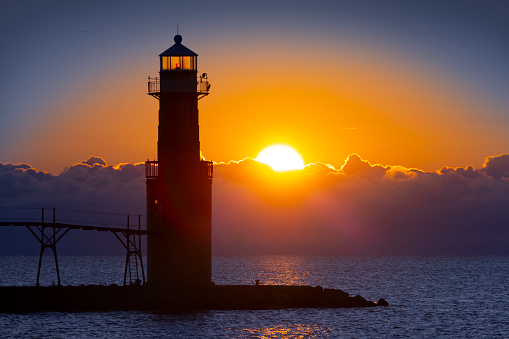 Scenic lighthouse with cormorant birds as sunrise breaks over a distant bank of fog on Lake Michigan. The light of night gives way to the light of day.