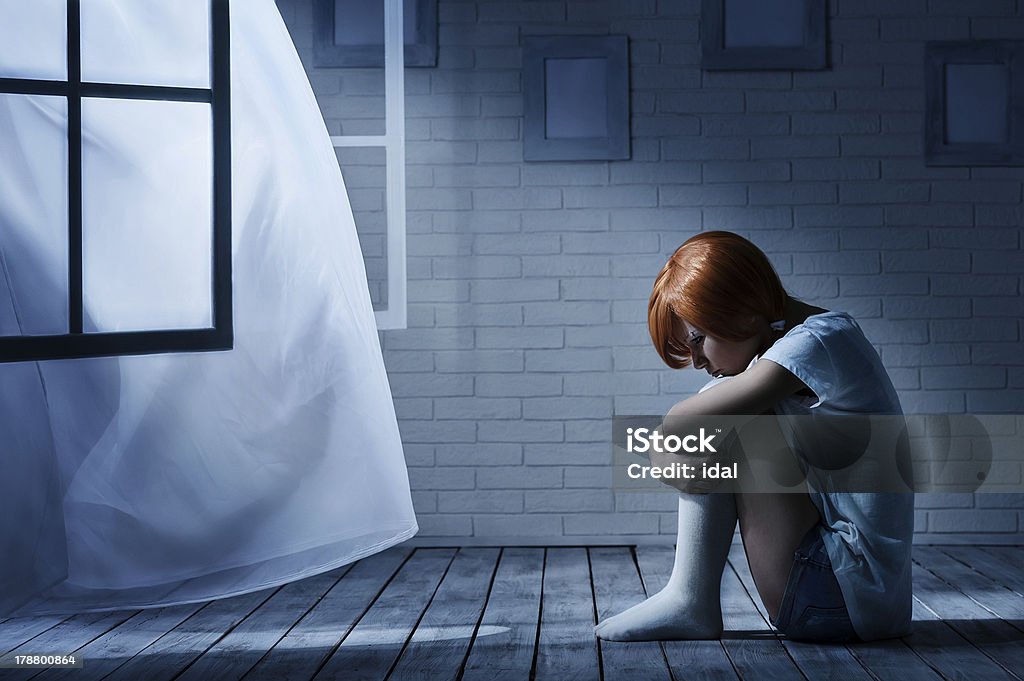 Lonely girl in a dark room Lonely girl sits in an empty dark room opposite the window Adult Stock Photo