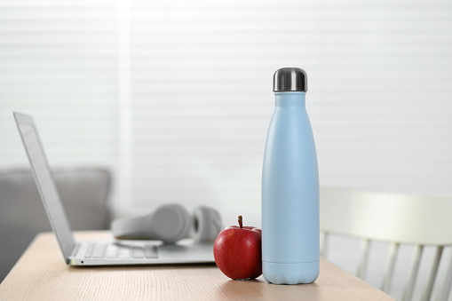 Stylish thermo bottle, apple and laptop on wooden table at home. Space for text