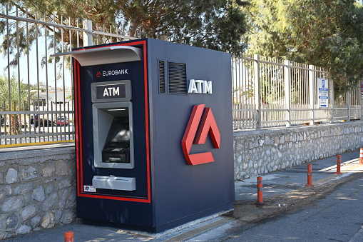 Heraklion, Greece 09 13 2023: Blue ATM machine of Eurobank is standing alone in the area of passenger seaport. Behind is metal fence and small stone wall.