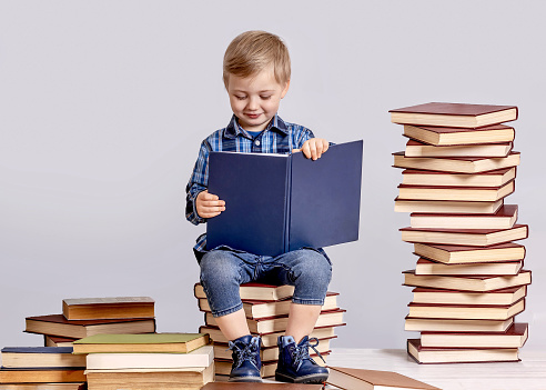 A three-year-old boy is reading a book while sitting on a stack of books. He smiles sweetly at the book. The concept of early childhood development.