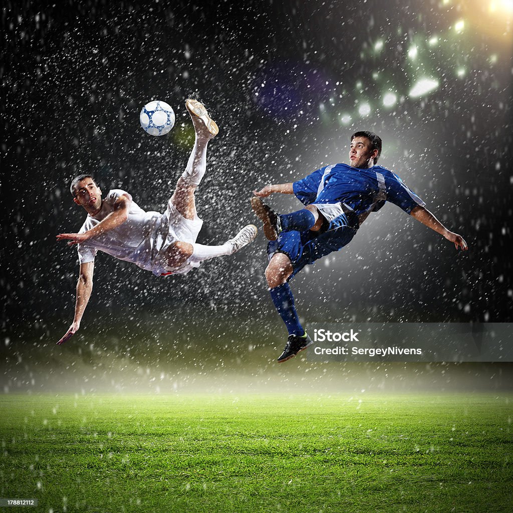 two football players striking the ball two football players in jump to strike the ball at the stadium under rain Above Stock Photo