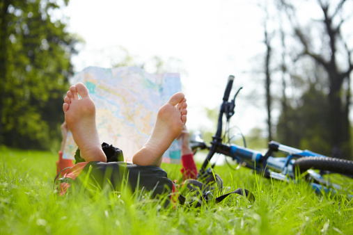 Girl cyclist reads a map lying barefoot on green grass outdoors in summer park. Enjoying relaxation