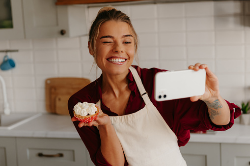 Joyful female baker using smart phone while making selfie with freshly made muffin on the kitchen