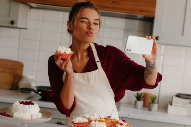istock Playful female baker using smart phone while making selfie with various cakes and muffins on the kitchen 1788152203