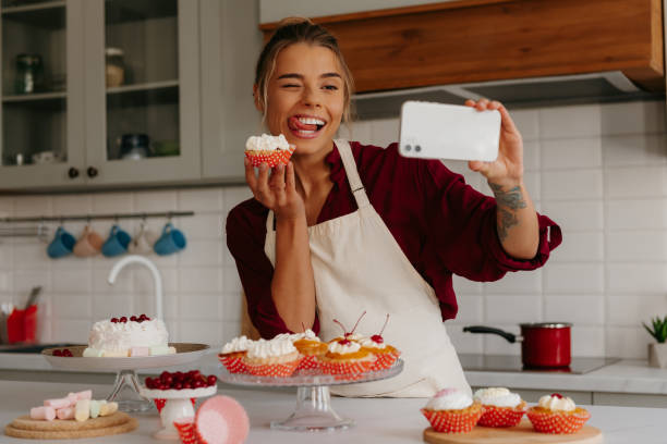 istock Joyful female baker using smart phone while making selfie with freshly made muffin on the kitchen 1788153219