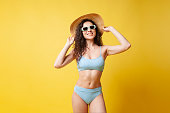 portrait of a young beautiful slim girl in a blue swimsuit and sunglasses on a yellow background, woman in a straw hat