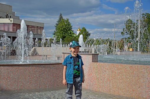 portrait of a cheerful boy during a walk near the fountain. the child smiles and enjoys a good day and a great mood. active lifestyle and walks in the fresh air. streams of water in the background