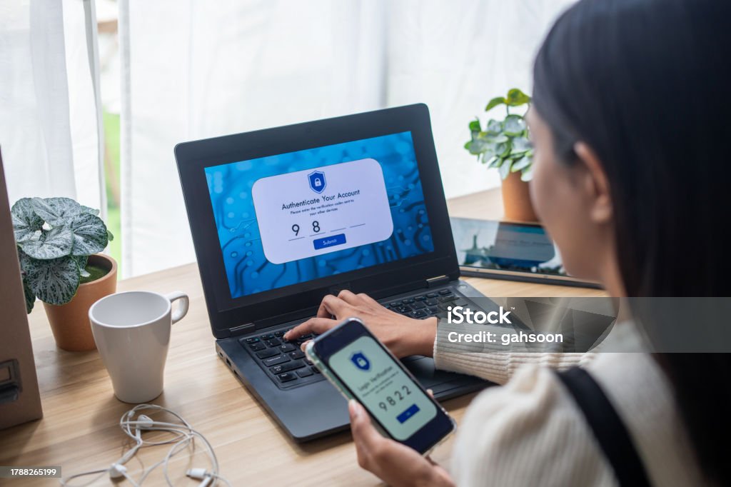 Two-Factor Authentication (2FA) security login in securely to her laptop over the shoulder shot of woman using smart phone with Two-Factor Authentication (2FA) security while logging to laptop. Privacy protection, internet and mobile security Digital Authentication Stock Photo