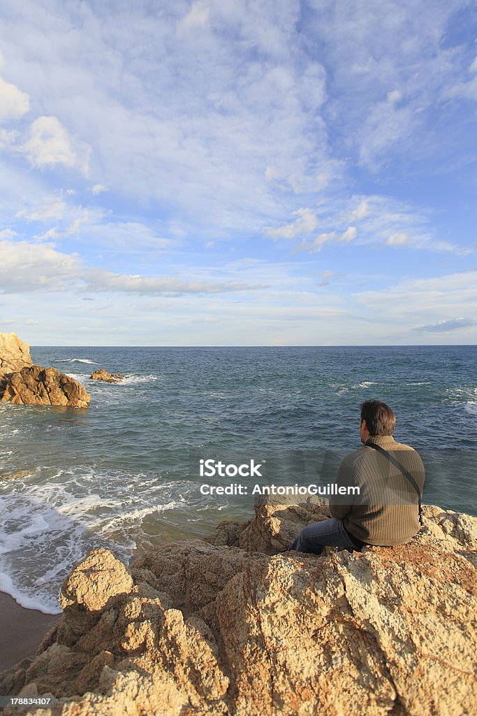 Hombre sentado en una roca en la playa mirando paisaje - Foto de stock de Actividades recreativas libre de derechos