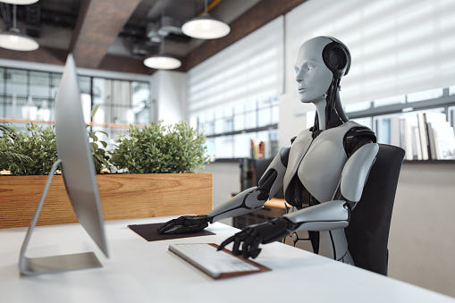 A futuristic humanoid robot uses a computer in a contemporary office.