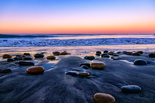 This is a beautiful sunrise photo taken early in the morning at moonlight beach California with rocks in the colorful magenta orange sand reflection of the sun asthe waves lap against the stony shore.