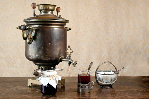 Old, samovar at the table. Glass of tea in glass holder. Retro sugar bowl with teaspoon. Jar with homemade preserves.