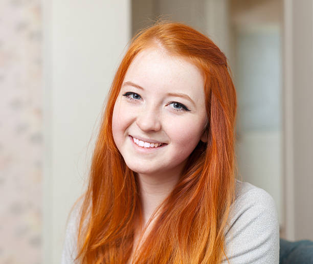 A cheerful woman with curly red hair leans forward, resting her chin on her hands, exuding a warm and happy ambiance in a bright indoor setting.