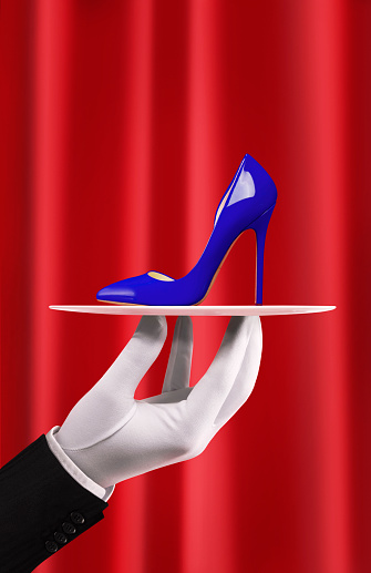 Butler with gloves holding and serving a chic women shoe with high heels in front of a red curtain