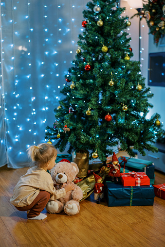 Little girl with a teddy bear is squatting near a Christmas tree with gifts underneath