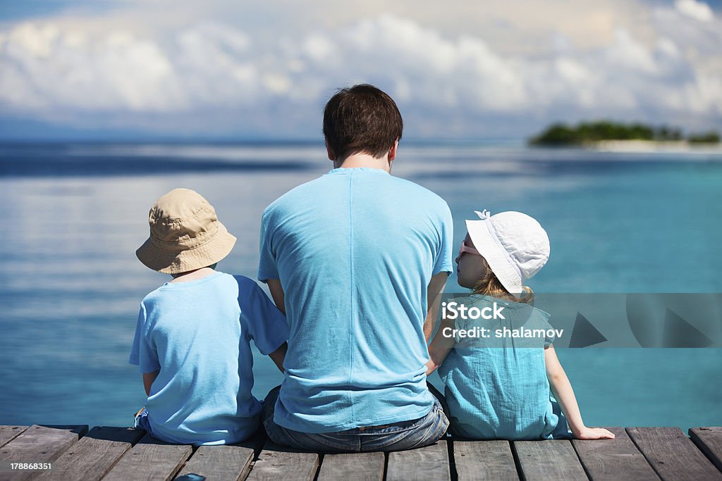 Father and kids enjoying ocean view Back view of father and kids sitting on wooden dock looking to ocean Adult Stock Photo