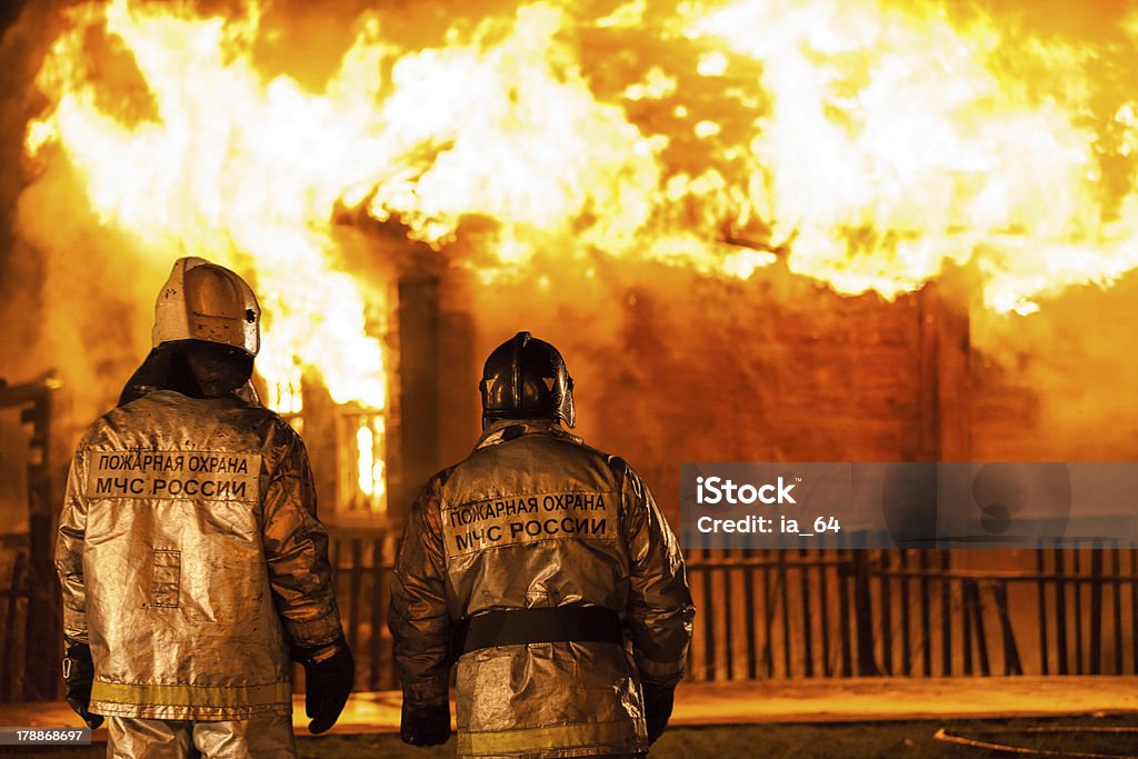 Firefighters at burning fire flame on wooden house roof Arson or nature disaster - firefighters at burning fire flame on wooden house roof. Inscription on back says they are workers of fire prevention department of Russian rescue service. Building Exterior Stock Photo
