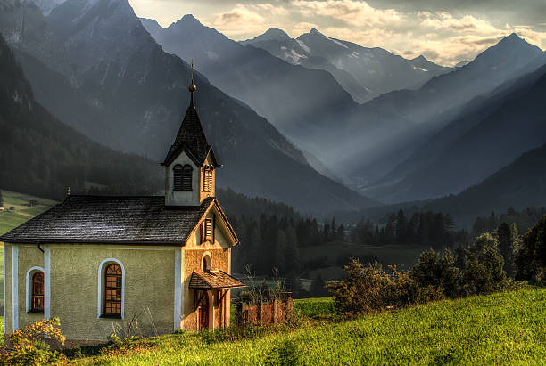 The chapel Maria Königin stands on a grassy hill near Lautersee in Upper Bavaria, Germany. Framed by tall trees and set against the dramatic peaks of the Karwendel Mountains under a cloudy sky, this scenic alpine location captures Bavarian culture, religion, and natural beauty in harmony.