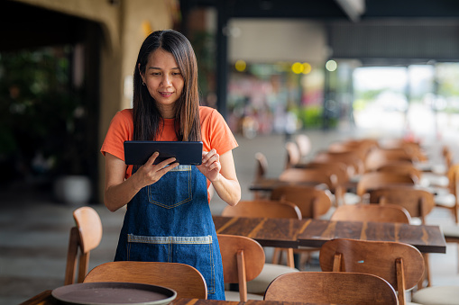 Portrait of Asian female smiling cafe owner holding digital tablet and wearing apron while standing at side walk cafe restaurant coffee shop  - Sustainability in small business,