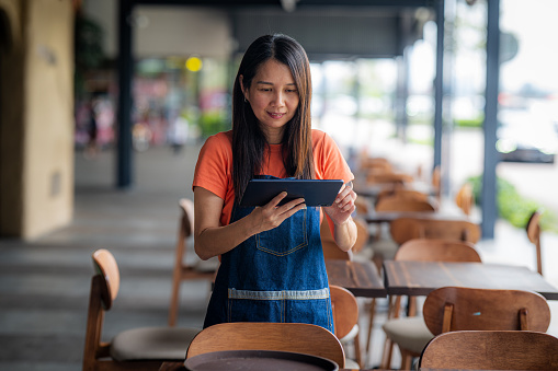Portrait of Asian female smiling cafe owner holding digital tablet and wearing apron while standing at side walk cafe restaurant coffee shop  - Sustainability in small business,