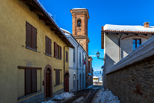 View of narrow street among old houses and belfry under blue sky in small town of Montelupo Albese in Piedmont, Northern Italy.
