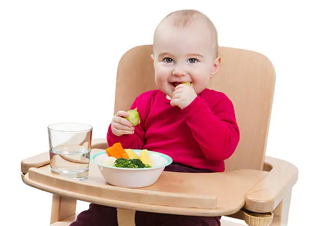 young child in red shirt eating vegetables in wooden chair.