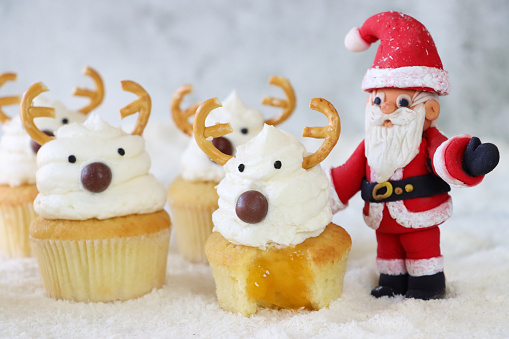 Stock photo showing close-up view of a batch of freshly baked, homemade Christmas reindeer design cupcakes displayed besides a Santa Claus figurine on icing sugar snow. The cupcakes are topped with a butter icing swirl decorated with candy coated chocolate sweet nose and salted pretzel antlers. One of the cakes has a missing bite from it showing the apricot jam filling. Home baking concept.