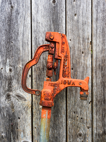 An old and weathered wooden barn wall serves as the backdrop for a rusty orange Waterford Iowa water hydrant pump. The contrast between the timeworn textures of the pump and the barn wall creates a rustic and nostalgic scene, echoing the history of rural life.
