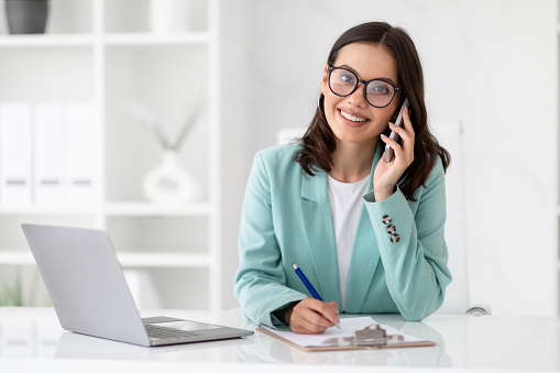 Smiling millennial caucasian lady secretary in suit and glasses calls by phone at workplace in light office interior. Business with device, communication with client, work, multitasking manager