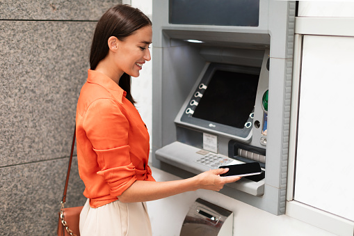 Young lady customer using smartphone to gain access to ATM, withdrawing money from account standing near bank machine on a street outside, side view shot. Financial safety and transactions