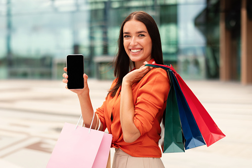 Shopping Application. Happy Customer Lady Showing Smart Phone With Blank Empty Screen For Mockup Holding Shopper Bag Standing Near Modern Mall Outdoors, Holding Cellphone Outside