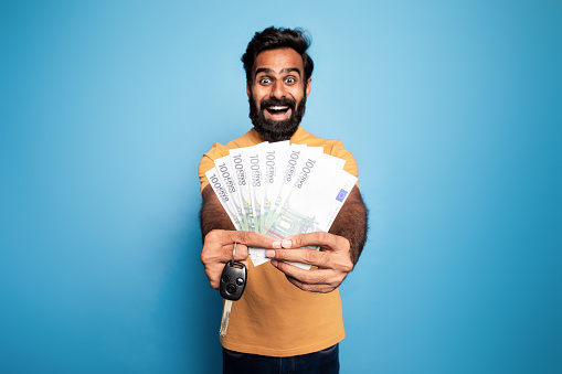 Make dream come true. Portrait happy indian man showing new car key and money cash dollars in hand, buying automobile, standing on blue studio background. Auto loan concept