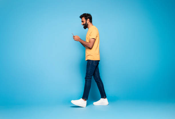 istock Indian guy in casual outfit walking with smartphone and smiling on blue studio background, full length, side view 1788998015