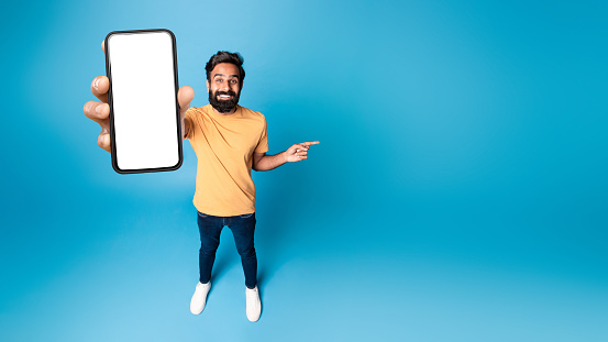 Mobile offer. Happy indian man showing phone with blank screen and pointing finger aside at free space on blue studio background, panorama, mockup. Choose this application