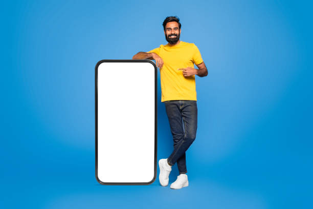 istock Indian man standing by huge smartphone with white blank screen, pointing at advertisement, blue background, mockup 1788998076