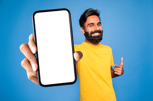 Online ad. Smiling middle aged indian man pointing at blank smartphone in his hand, standing over blue background, mockup. Happy hindu male recommending app or website, collage