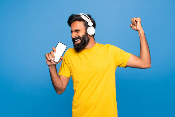 istock Happy indian man in wireless headphones singing at smartphone with empty screen in imaginary microphone, blue background 1788998216