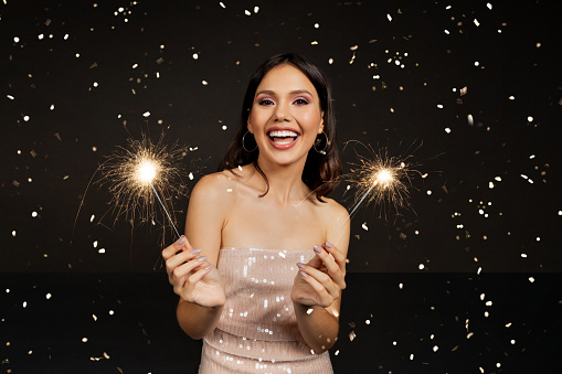 Excited beautiful young woman wearing nice dress and makeup hold sparklers, enjoying party, celebrating anniversary or New Year, posing alobe over black background with falling confetti, Free Space