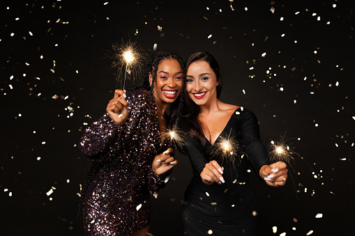 Happy multiethnic girlfriends two young women wearing nice elegant outfits holding bengal lights, laughing among confetti sparkles, celebrating New Year 2024 together, isolated on black background