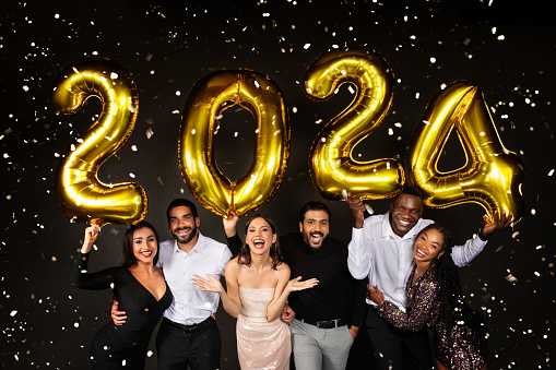 Happy multiracial men and women three loving millennial couples wearing nice outfits celebrating new year 2024 together on black background, holding golden number baloons and smiling at camera