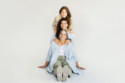 Glad caucasian different women hugging, have fun, enjoy love and spare time, sit on floor on white wall background. Family relationships, grandmother, mom and daughter, full length