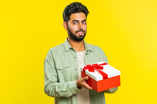 Positive smiling Indian man presenting birthday gift box stretches out hands, offer wrapped present career bonus, celebrating party, promotion discount sale. Arabian guy isolated on yellow background