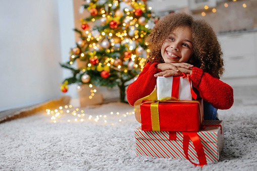 A black girl in a red sweater with lots of gifts for Christmas. A happy child near the Christmas Tree in his room.
