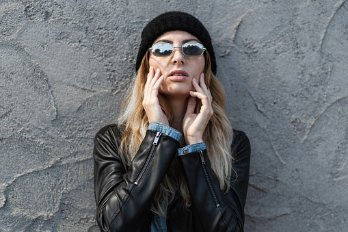 Woman in a chic leather jacket and beanie posing confidently, her sunglasses reflecting the city vibe around her.