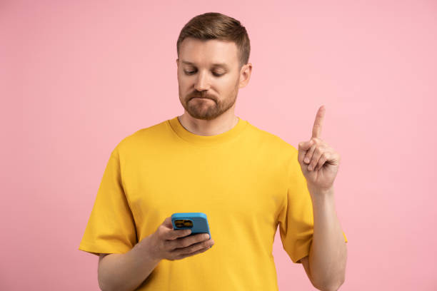 istock Sad upset man pointing index finger up looking in smartphone on pink background. 1789112097
