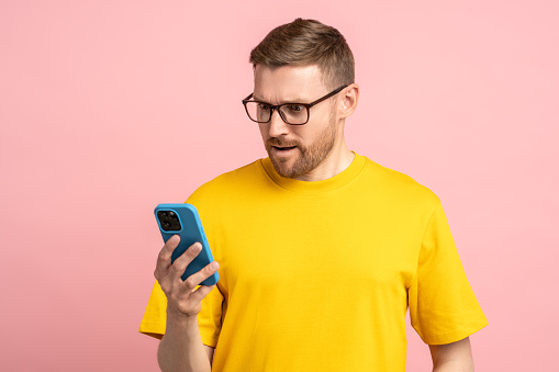 Shocked surprised astonished man in glasses holding smartphone, reading news, message, checking online lottery results with amazed stunned look. Middle-aged puzzled man isolated on pink background