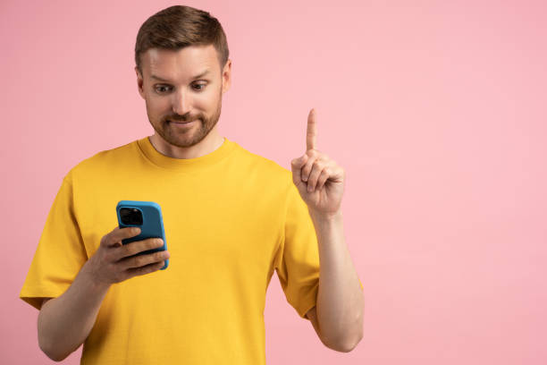 istock Interested man shows finger sign Idea eureka looks on smartphone screen on pink studio background 1789112102