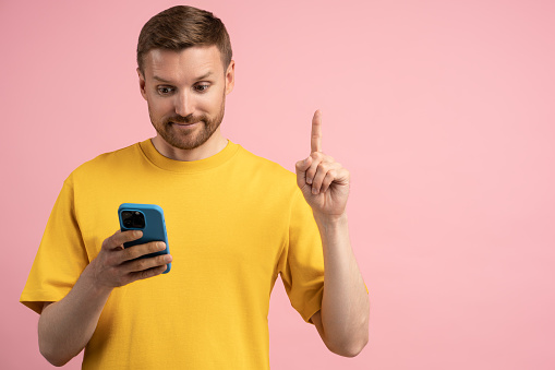 Interested man shows finger sign Idea receive offer by message on mobile phone on pink studio background. Smiling guy stands with attention gesture extends index finger up looking on cellphone screen